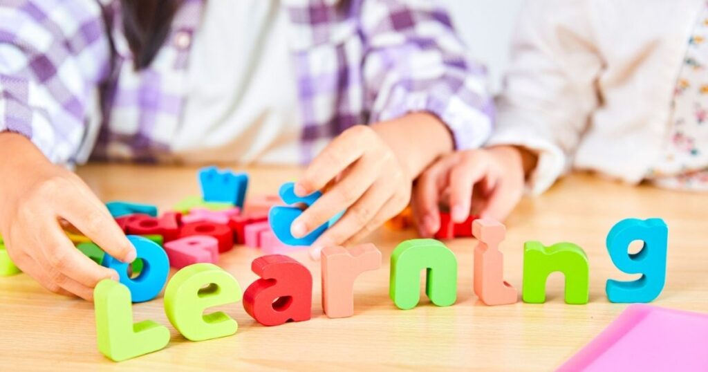 Two children arrange colorful wooden letters to spell 'learning' on a wooden table.