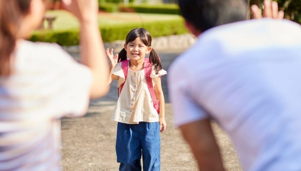 Livelihood Parenting -What a Live Phonics Course for Mothers in Hinglish Looks Like - Livelihood Parenting by Honey Maheshwari Young girl with a pink backpack waves hello while standing on a park path, flanked by adults reaching out to her.
