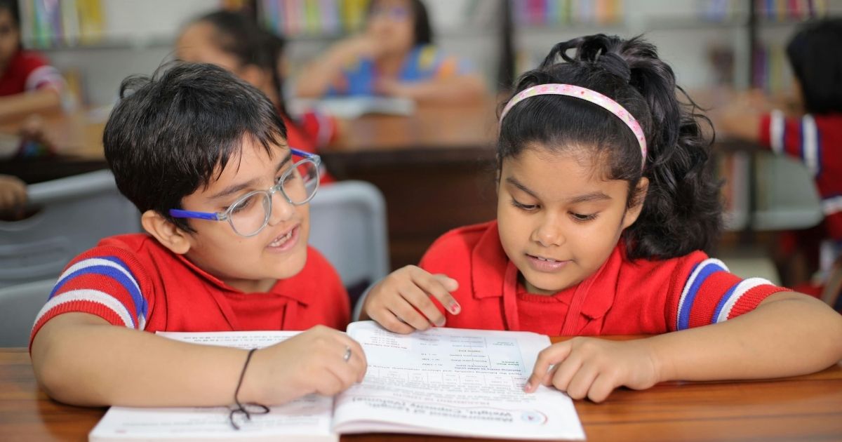Livelihood Parenting -Decodable Books for Kids and Why They Beat Pretty Picture Books at First - 2 - Livelihood Parenting by Honey Maheshwari - Livelihood Parenting by Honey Maheshwari Two young students in red uniforms sit at a wooden desk, reading and pointing at a workbook together in a classroom with shelves in the background.